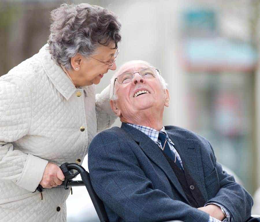 Smiling elderly couple, wofe pushing husband in wheen chair.