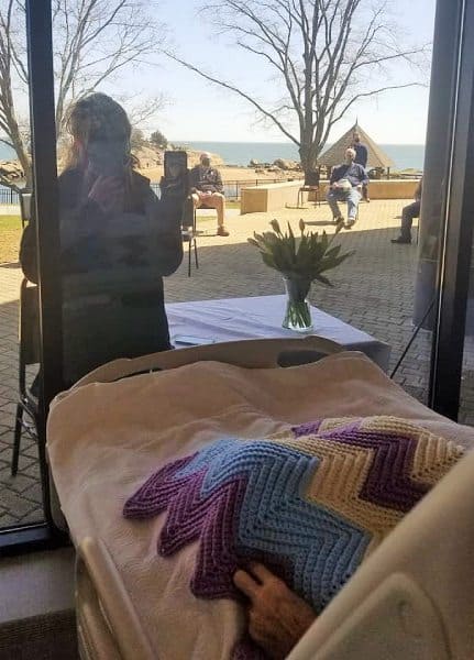 Connecticut Hospice patient in bed facing the waterfront, with family member taking video of her through window, while more family sits apart. 
