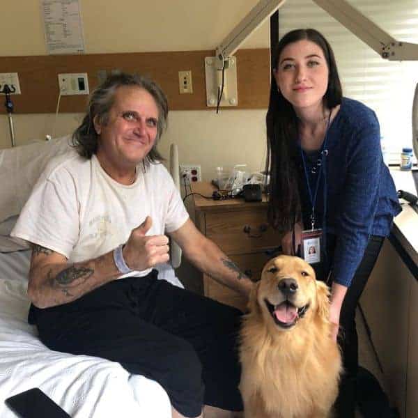 Patient sitting on edge of bed stroking therapy dog, smiling and giving thumbs up