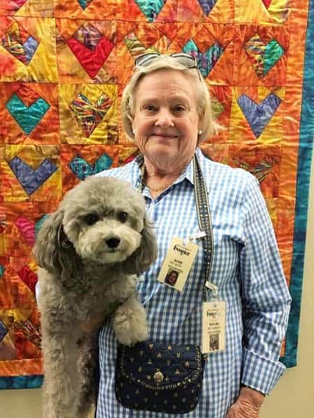 Proud pet therapy volunteer stands in front of colorful quilt, holding therapy dog under one arm