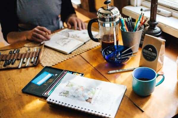 Man draws in sketchbook at table with art supplies and coffee nearby

