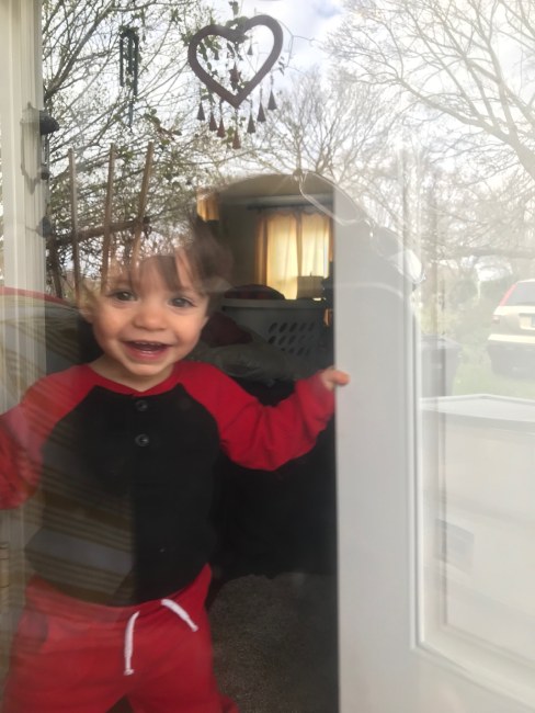 Toddler smiles widely as he holds door open to greet socially distanced grandparent through window