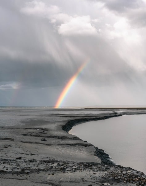 rainbow shines down to edge of water through grey clouds
