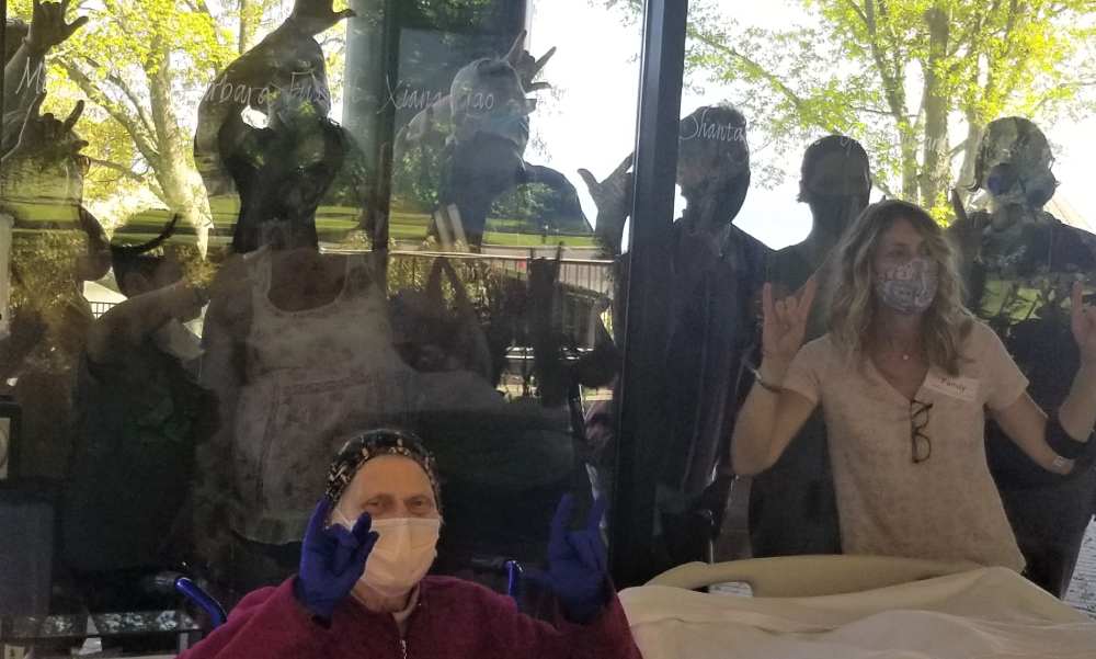Wife of patient sits next to bed while all family members cluster on other side of glass window and make "I Love You" sign with fingers
