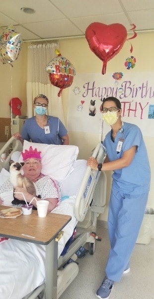Hospice Social work  and Arts Program staff members stand next to patient's bed with balloons and birthday banner hanging behind.