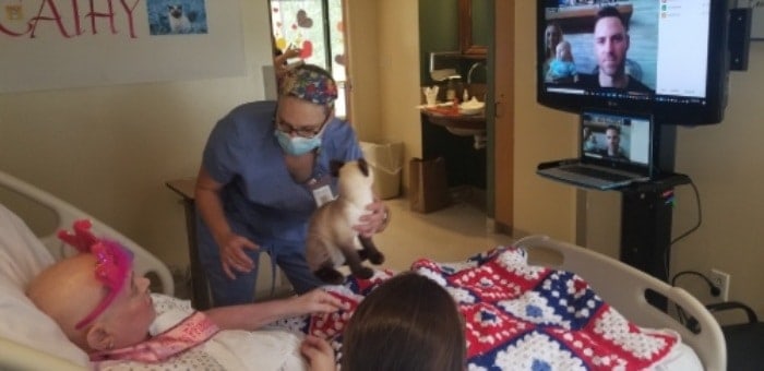 Hospice patient's family members are pictured on tv screen facing hospice patient's bed while staff 