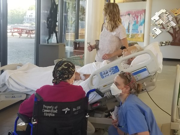 Sleeping patient lies in bed in Connecticut Hospice lobby with wife in wheelchair next to him, daughter standing next to bed, and staff social worker crouches down to speak to wife