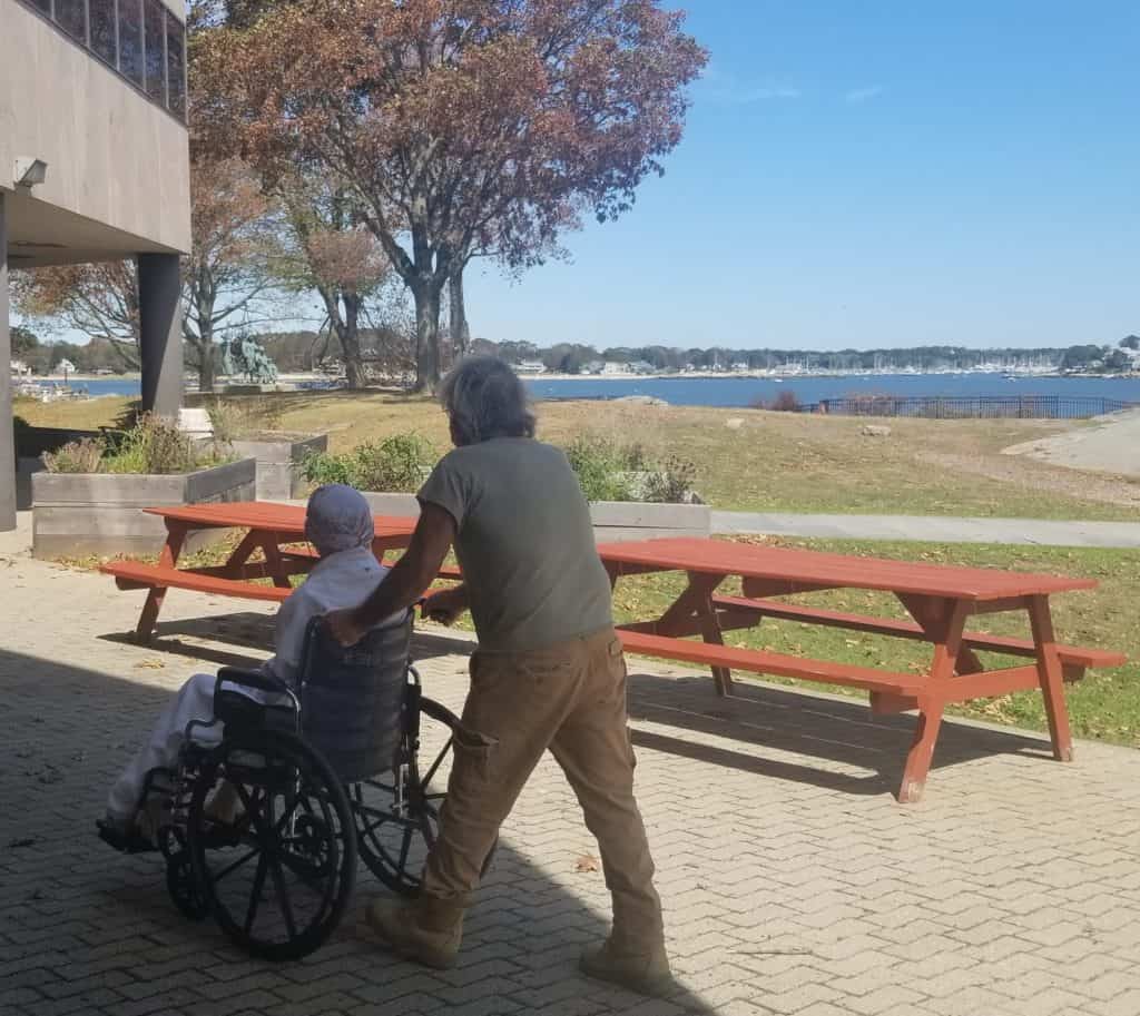 male volunteer pushing female hospice patient in wheelchair on outside grounds of Ct Hospice in-patient facility located on shoreline with views of blue water and skies.  