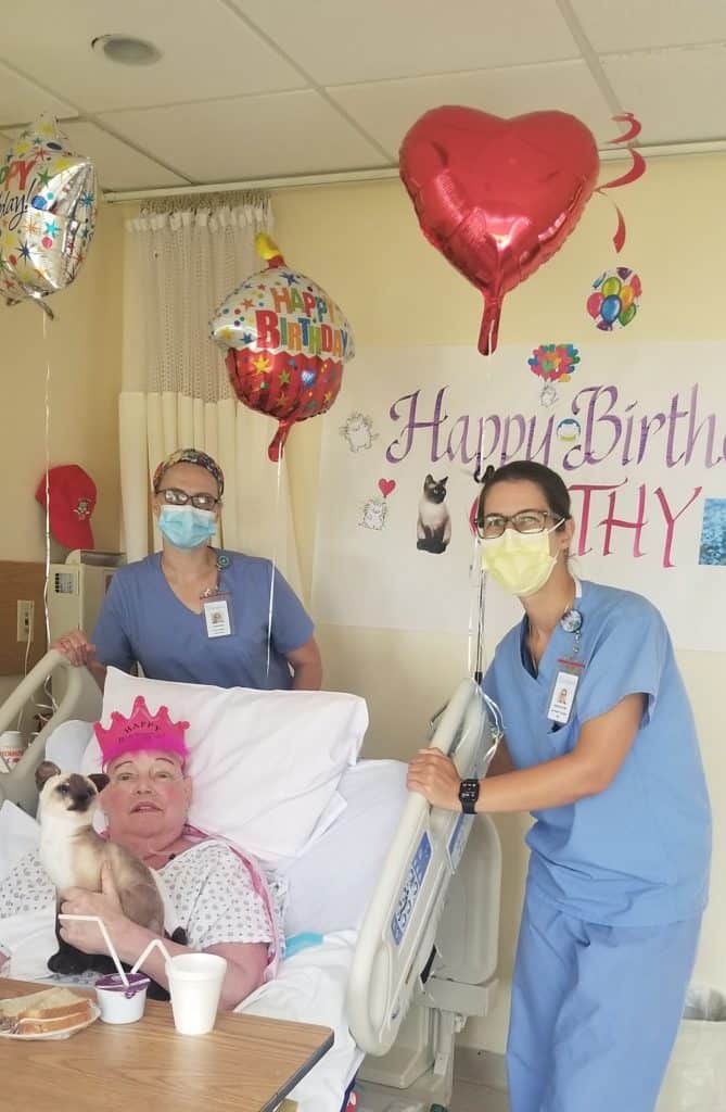Female patient in hospital bed wearing pink crown and holding stuffed cat celebrating birthday with two hospital staff members