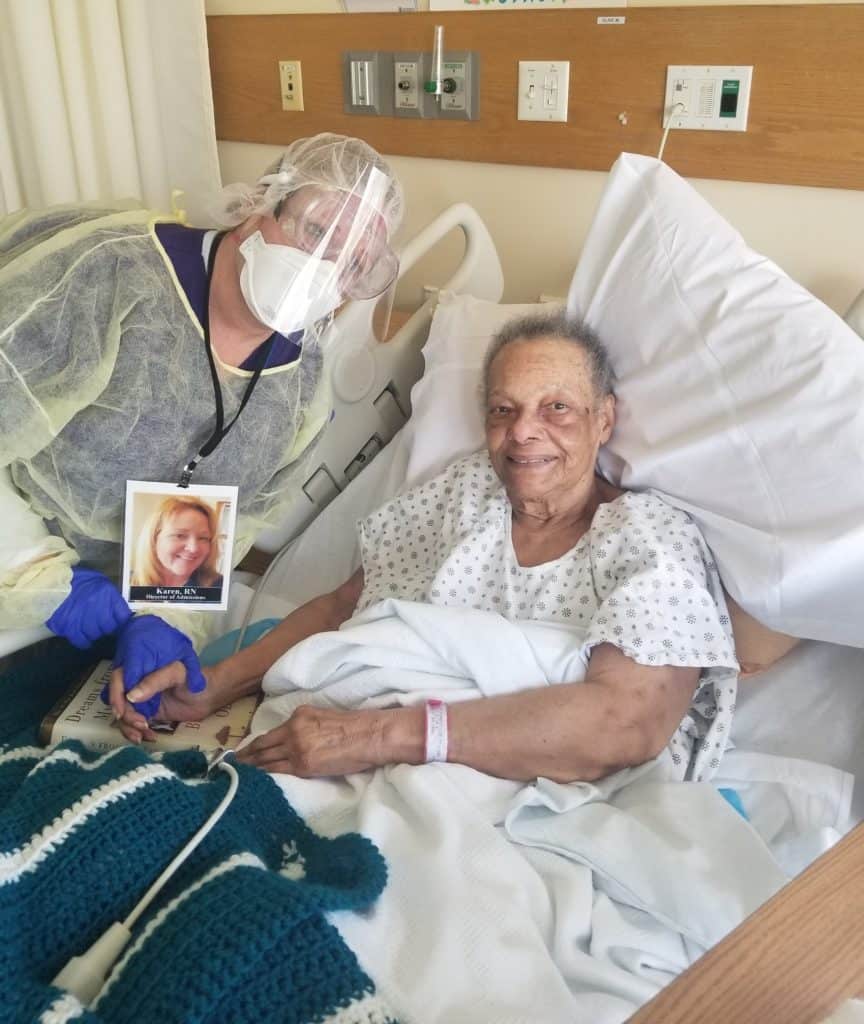 Hospice female patient being visited at bedside by hospice nurse wearing full PPE (yellow gown, yellow head protection, purple gloves, clear eye goggles, and mask)  