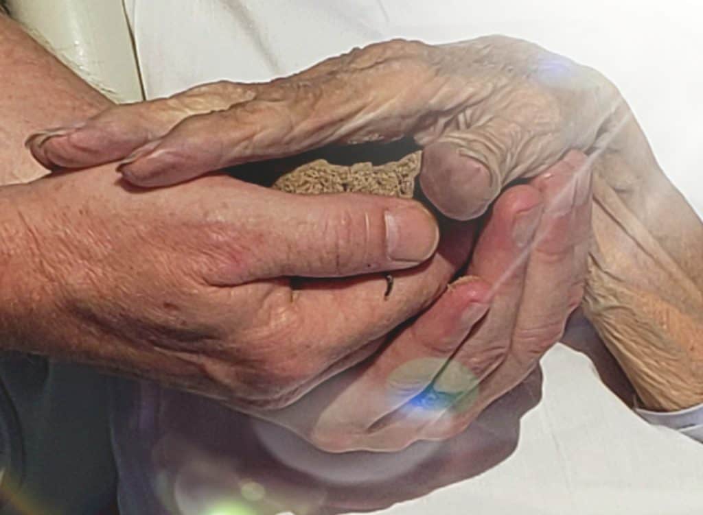 several hands supporting a hospice patient's hands that are holding a mound of sand 
and all supported by a white glowing light