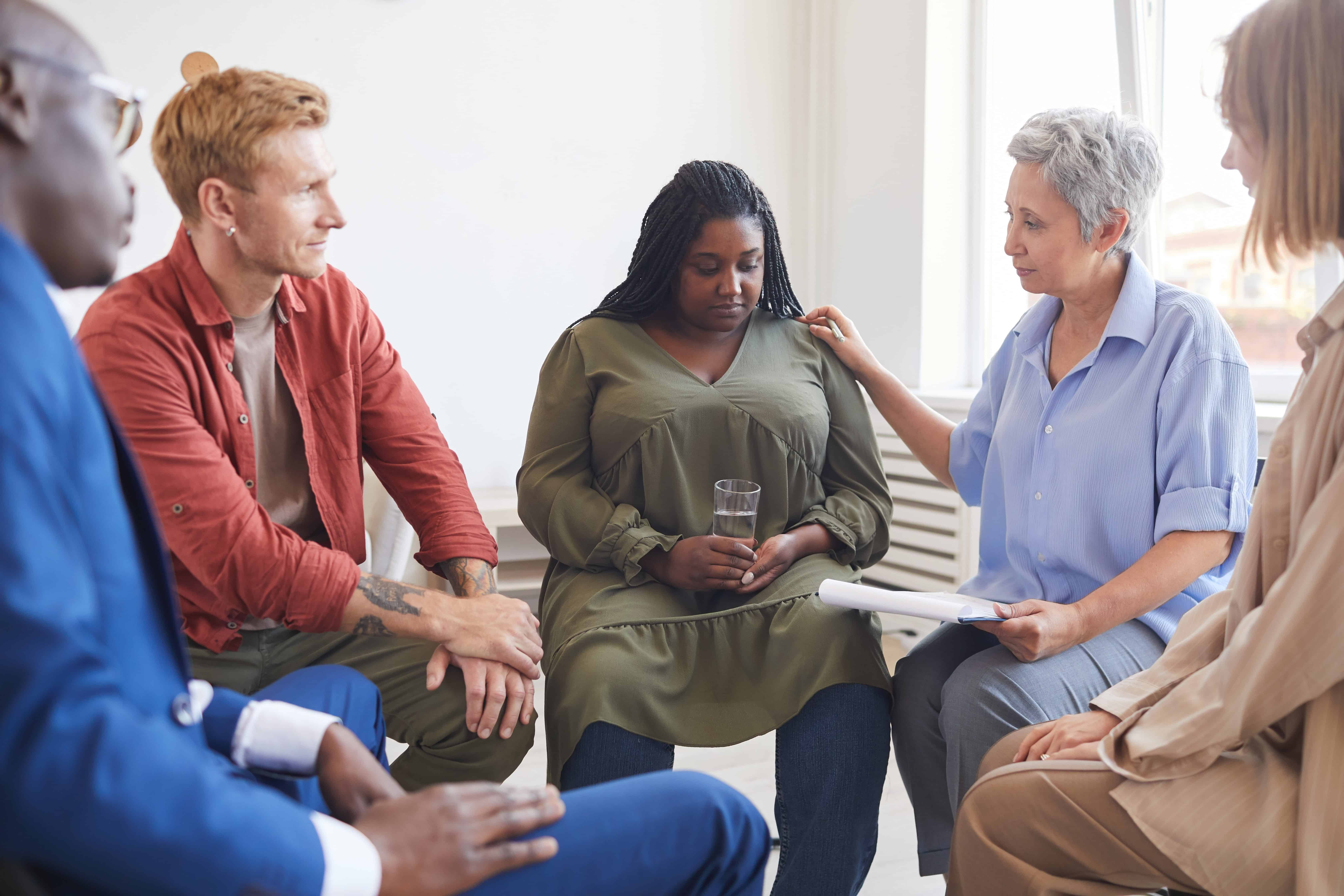 Portrait of young woman sharing struggles during support group meeting with people siting in circle and comforting her